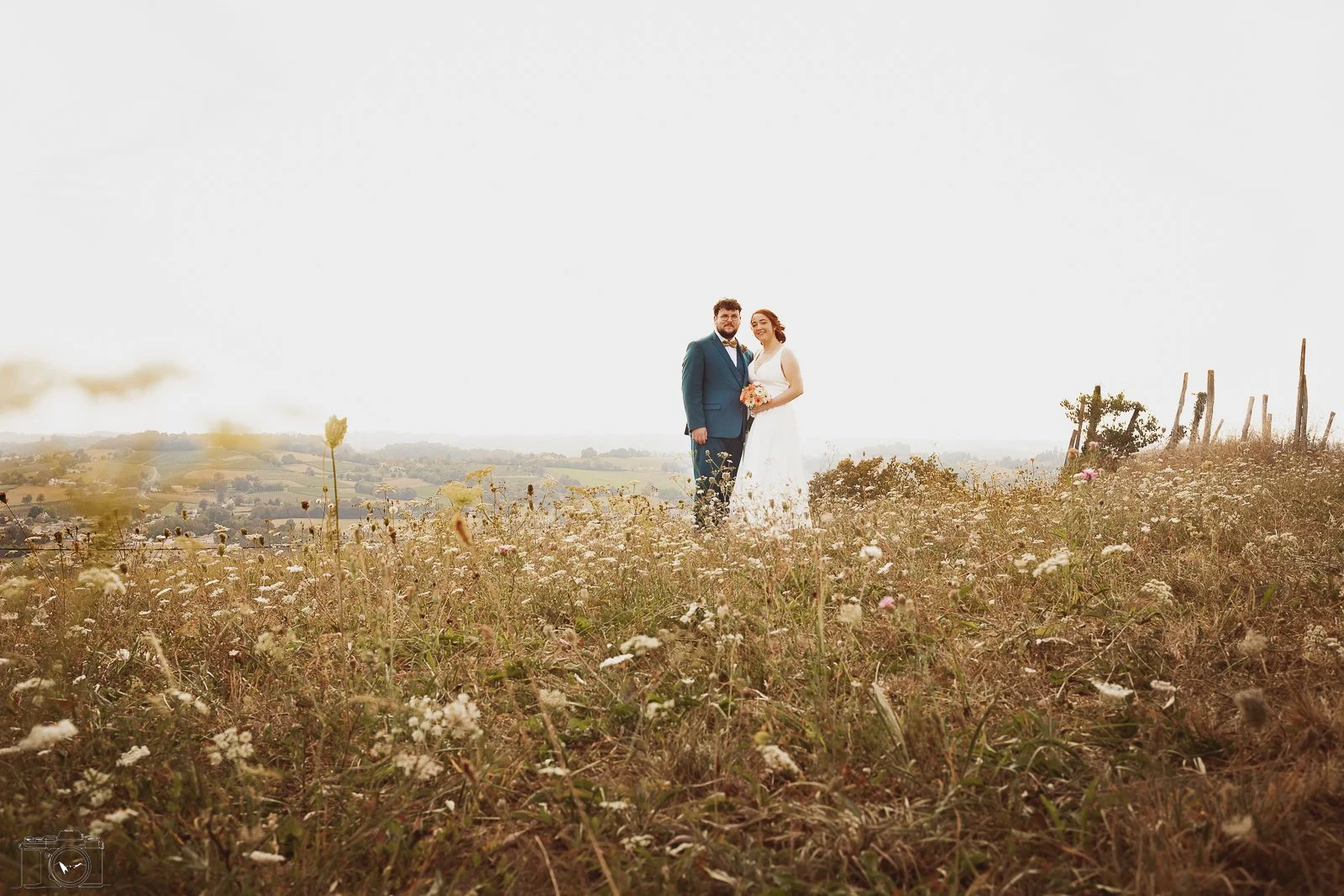 Un couple de mariés debout dans un champ de fleurs sauvages avec un paysage rural à l'arrière-plan, ciel clair.