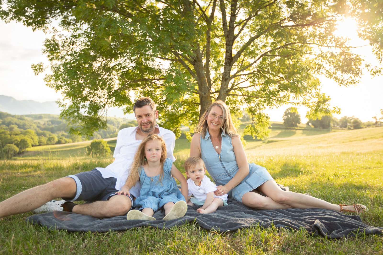 Famille de quatre personnes, deux adultes et deux enfants, assis sur une couverture dans un champ verdoyant avec un arbre en arrière-plan, sous un soleil couchant.