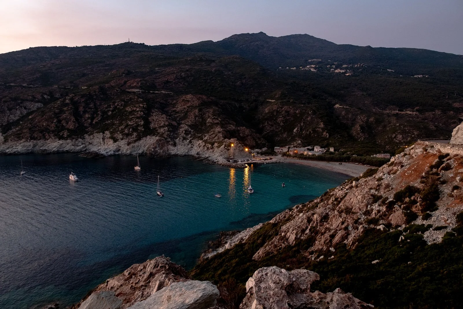 Vue nocturne d'une petite baie avec des voiliers au calme, entourée de collines rocheuses avec quelques maisons éclairées.