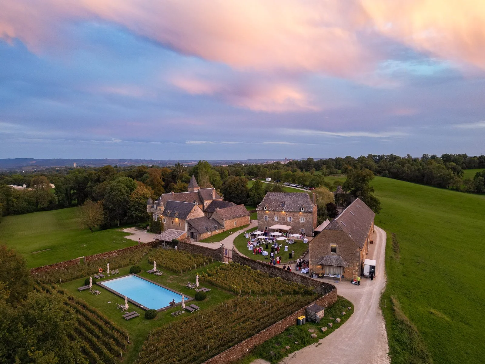 Un village rural avec des bâtiments en pierre, un jardin avec une piscine et une terrasse où se déroule un événement près d'une campagne verdoyante sous un ciel coloré au coucher du soleil.