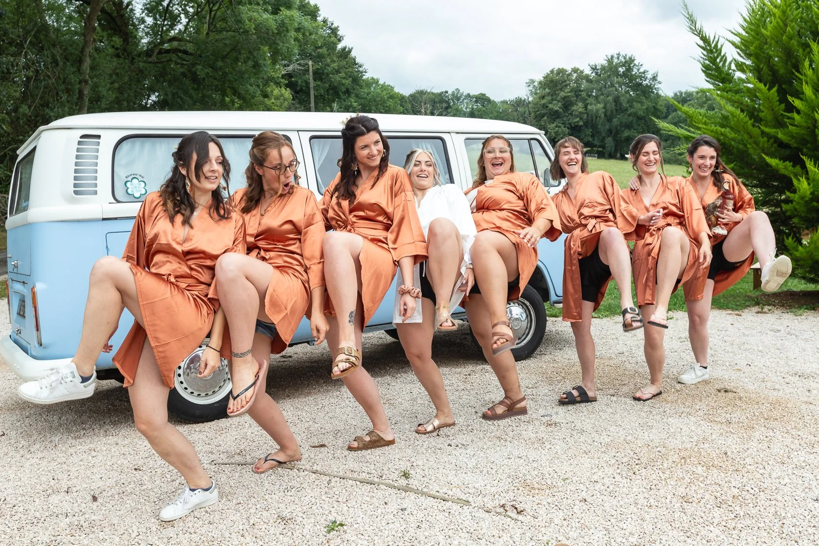 Groupe de huit femmes souriantes portant des robes de mariée en satin brun orangé, assises sur un van vintage bleu et blanc, dans une ambiance joyeuse en plein air avec des arbres verts en arrière-plan.