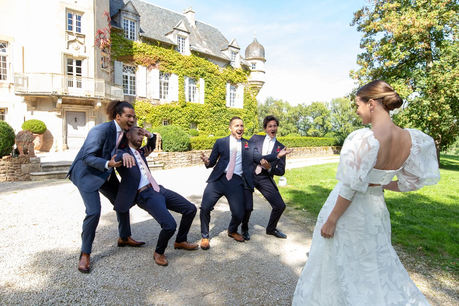 Un groupe d'hommes en costumes souriants et riant face à une femme en robe de mariée blanche devant une grande maison ancienne avec des jardins. C'est un mariage ou une séance photo de mariage.