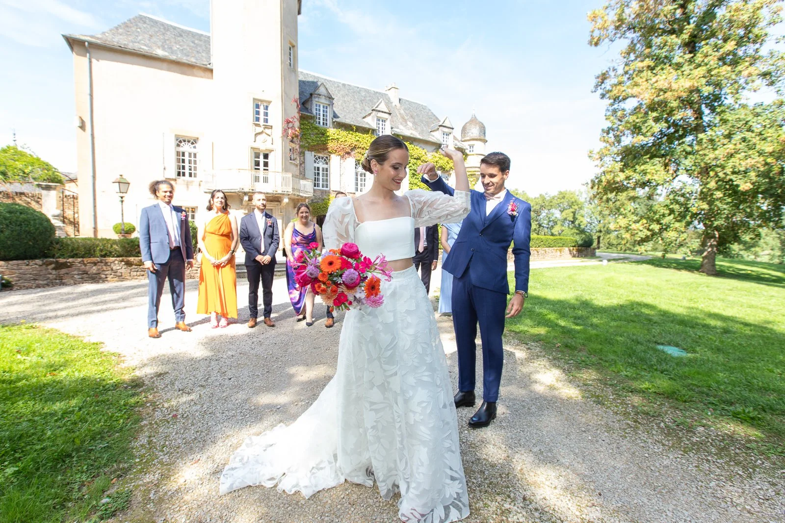 Un mariage en extérieur avec un couple marié au centre, la mariée en robe blanche tenant un bouquet coloré, entourée de leurs invités dans un jardin devant un château. La mariée montre ses biceps en souriant, tout le monde semble heureux.