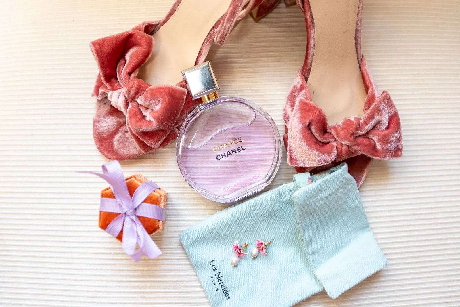 Une bouteille de parfum Chanel Chance entourée d'un collier en velours rose, d'une paire de boucles d'oreilles en or avec des perles, d'un sac en tissu vert pastel avec une fille en tissu orange avec un ruban violet.