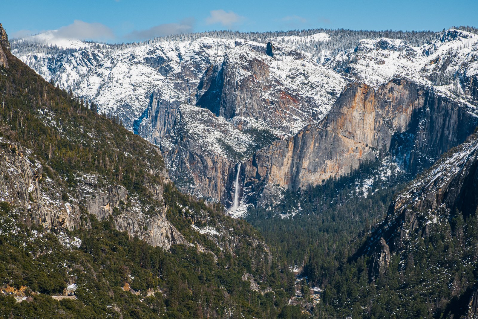 Paysage de montagne avec sommets enneigés, forêt dense en bas et cascade au centre.