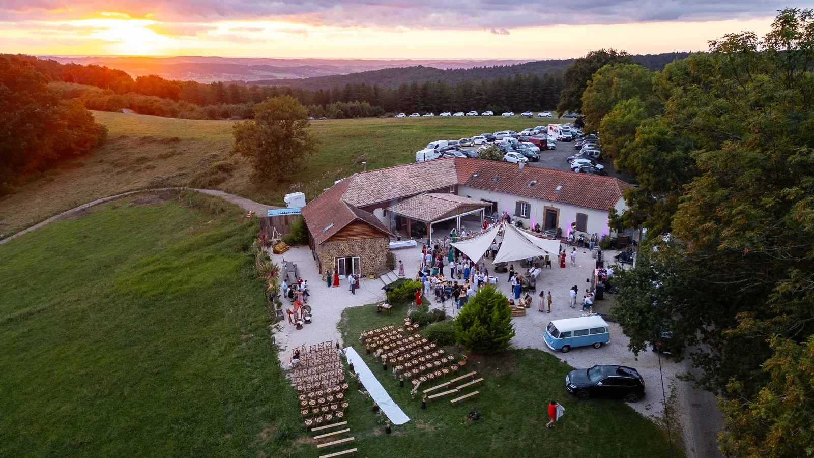Une réception ou fête en plein air avec de nombreux invités, une tente blanche, un bâtiment en pierre avec un toit en tuiles, des voitures garées, et un décor de campagne au coucher du soleil.