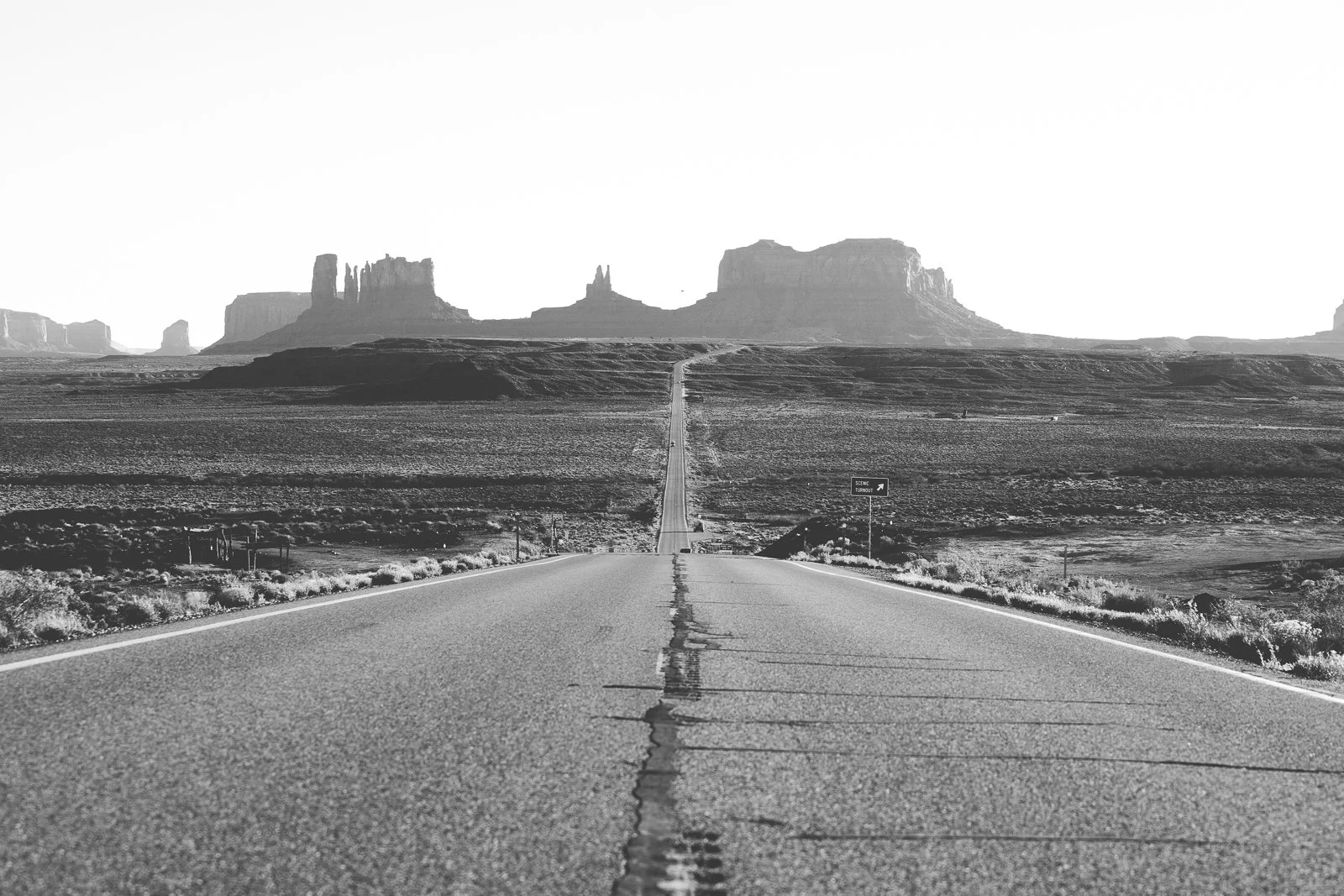 Une route droite dans un désert avec des formations rocheuses en arrière-plan, en noir et blanc.