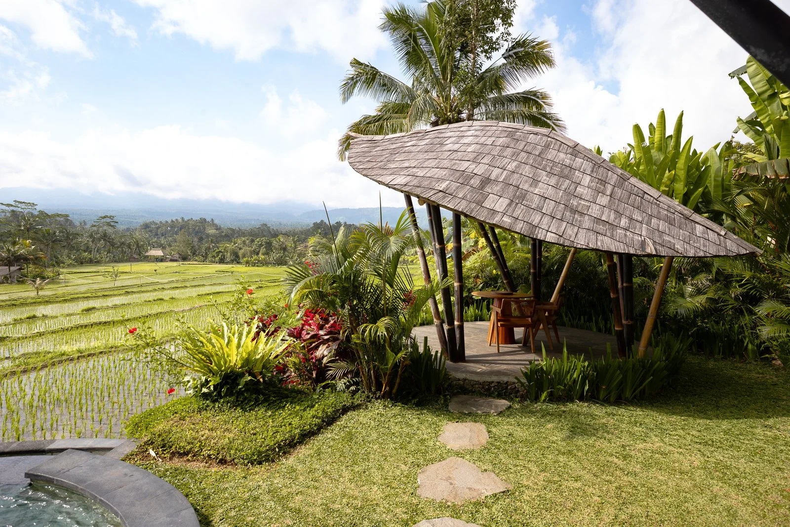 Pagode en bois avec toit en chaume dans un jardin tropical avec chaises et table, vue sur un paysage de rizières et montagnes au loin.