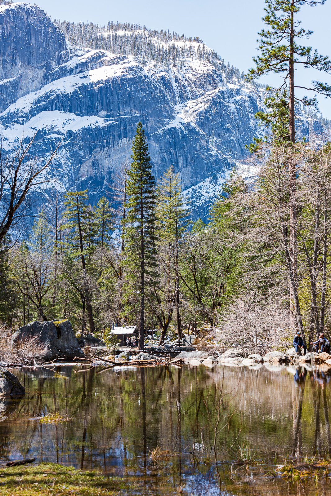 Montagnes enneigées, forêt de conifères verticaux, un lac calme avec des reflets d'arbres, quelques randonneurs assis sur des rochers à droite, ciel bleu clair.