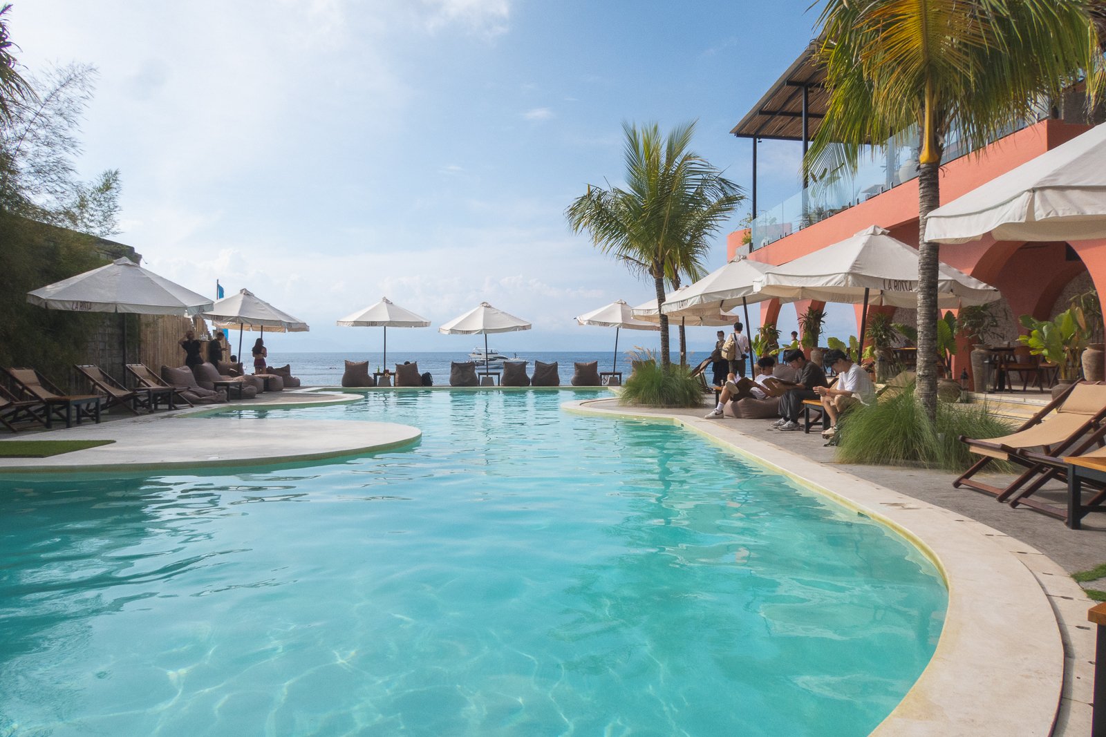 Piscine à débordement avec vue sur l'océan, parasols et chaises longues, personnes assises et se relaxant, bâtiment rose avec terrasse en haut, palmiers, ciel ensoleillé.