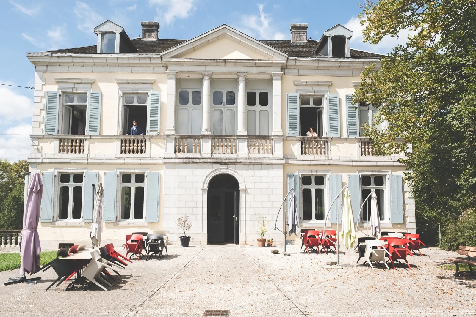 Villa classique avec balcons, fenêtres à volets bleus, et terrasse avec chaises et parasols, sous un ciel bleu avec quelques nuages.
