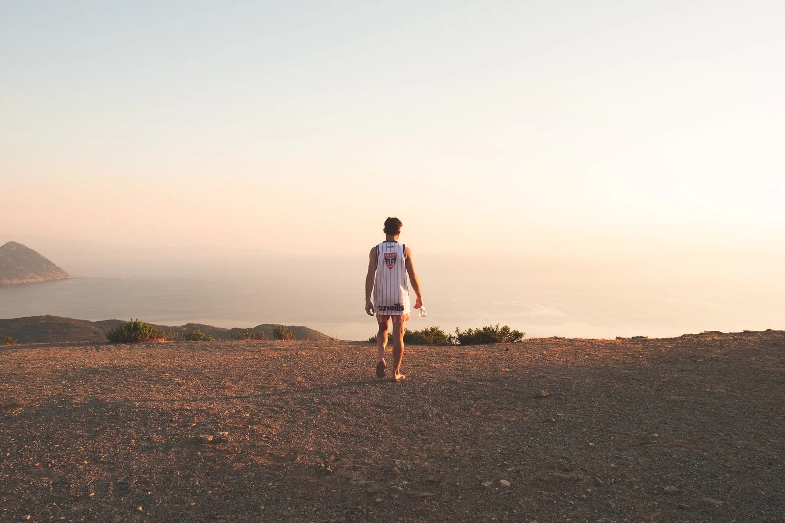 Un jeune homme en tenue de basket-ball avec un maillot blanc regarde vers l'horizon au coucher du soleil, sur un terrain dégagé avec vue sur la mer et des collines lointaines.