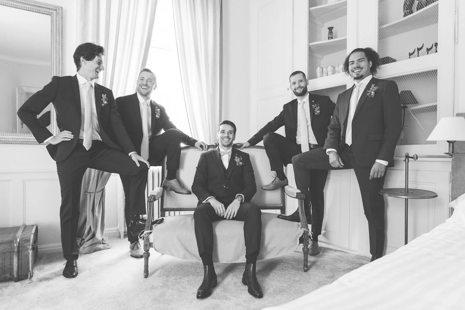 Groupe d'hommes en costume de mariage dans une pièce élégante, certains assis ou debout sur un canapé et meubles, tous souriants et posant pour une photo.
