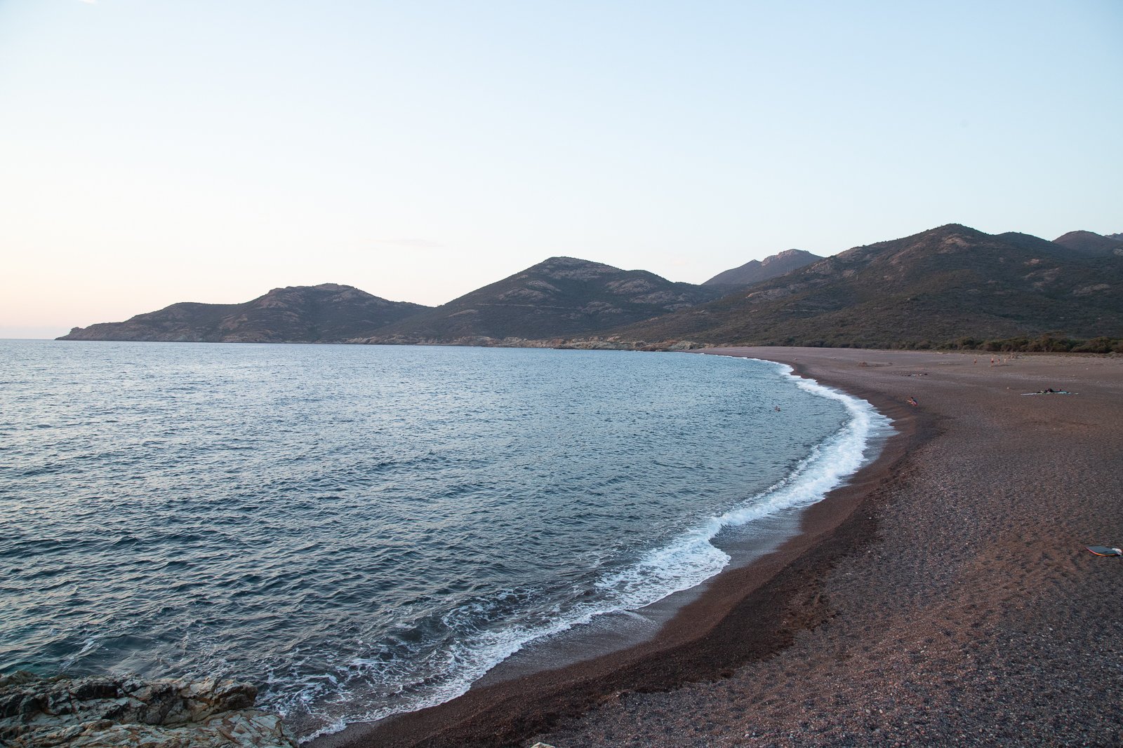 Plage de sable noir avec des montagnes en arrière-plan, eaux calmes et quelques personnes au loin
