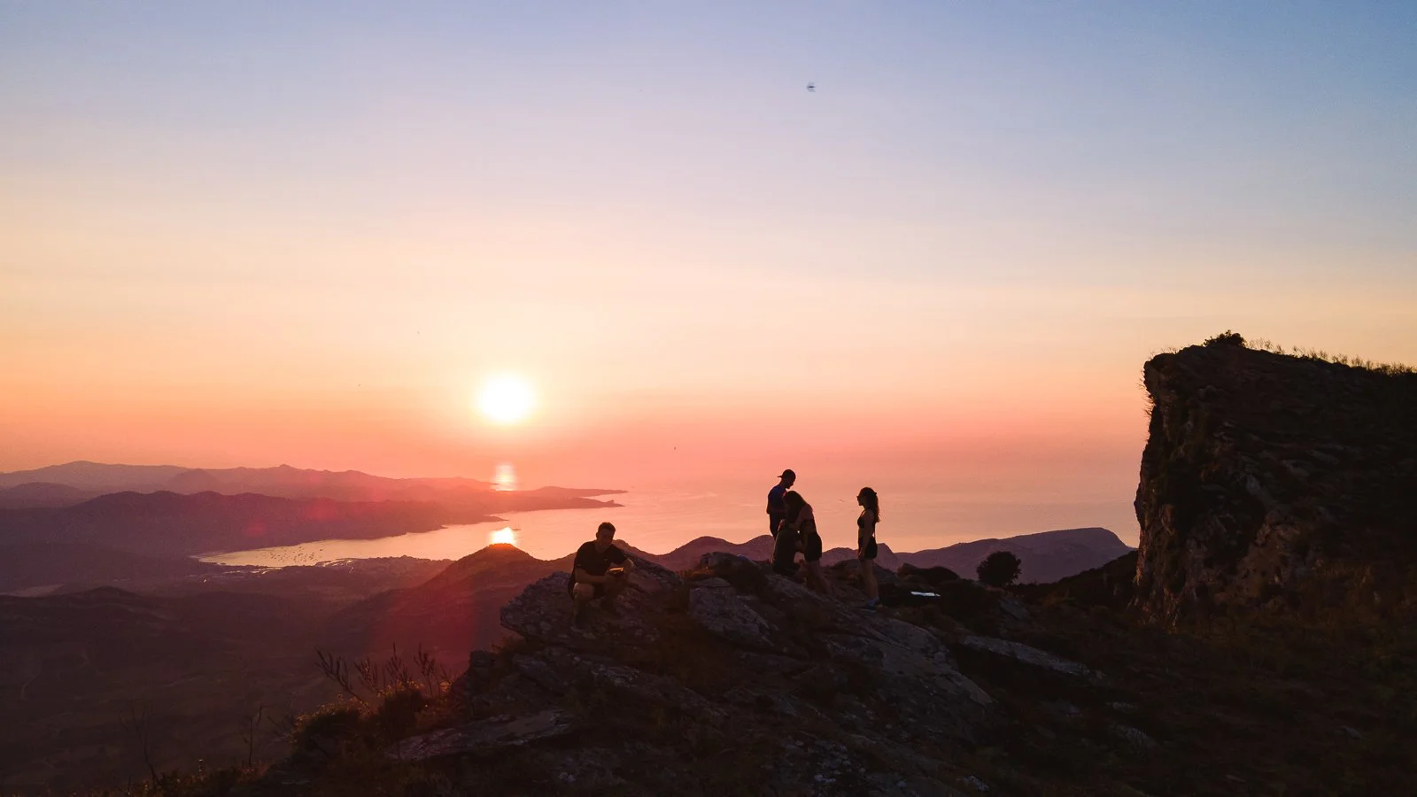 Groupe de personnes au sommet d'une montagne regardant le coucher de soleil sur un paysage côtier.