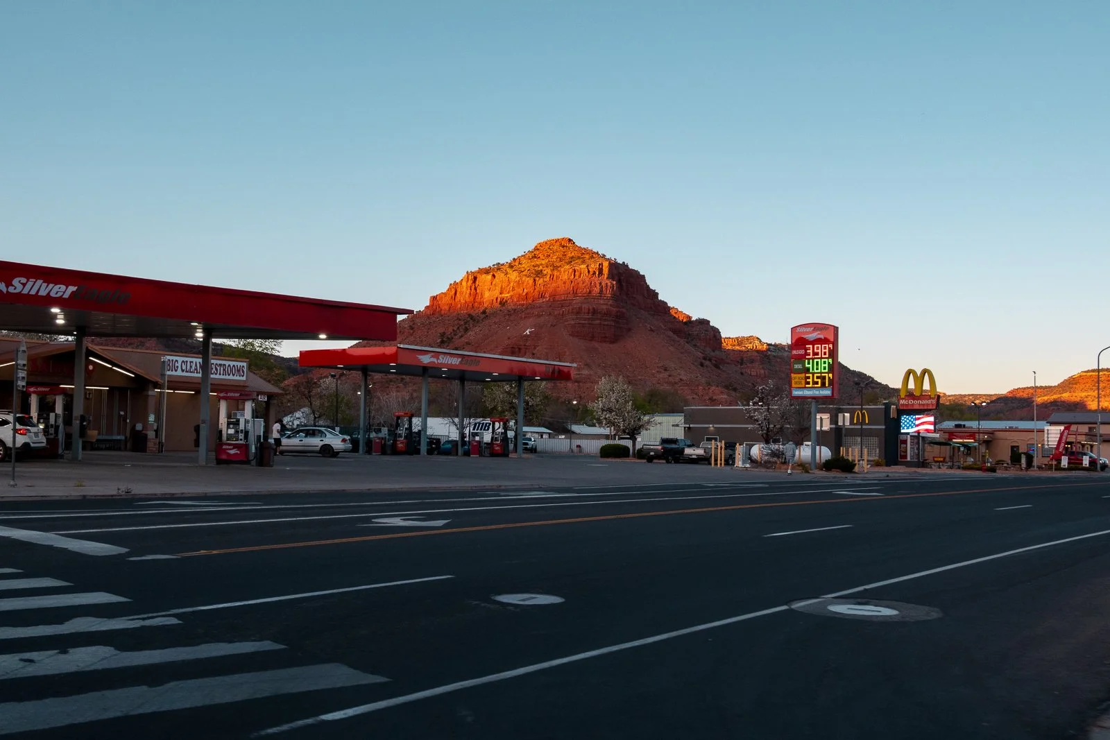 station-service en ville avec montagnes en fond, enseignes McDonald's, station d'essence Silver Eagle, et panneau de prix de carburant au coucher de soleil.