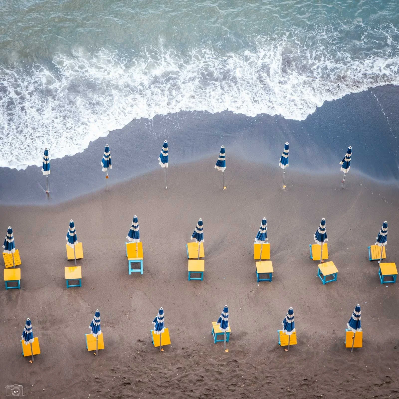 Plage vide avec des parasols bleus et jaunes alignés, les vagues de la mer arrivant sur le rivage