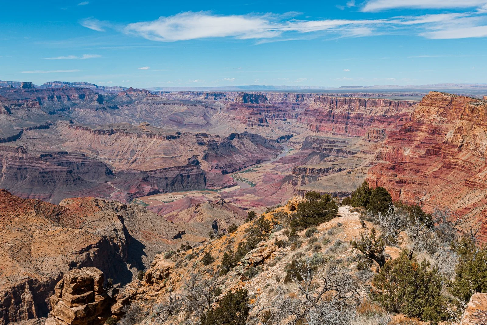 Vue panoramique du Grand Canyon avec ses falaises colorées, le fleuve Colorado en bas, et un ciel bleu avec quelques nuages.