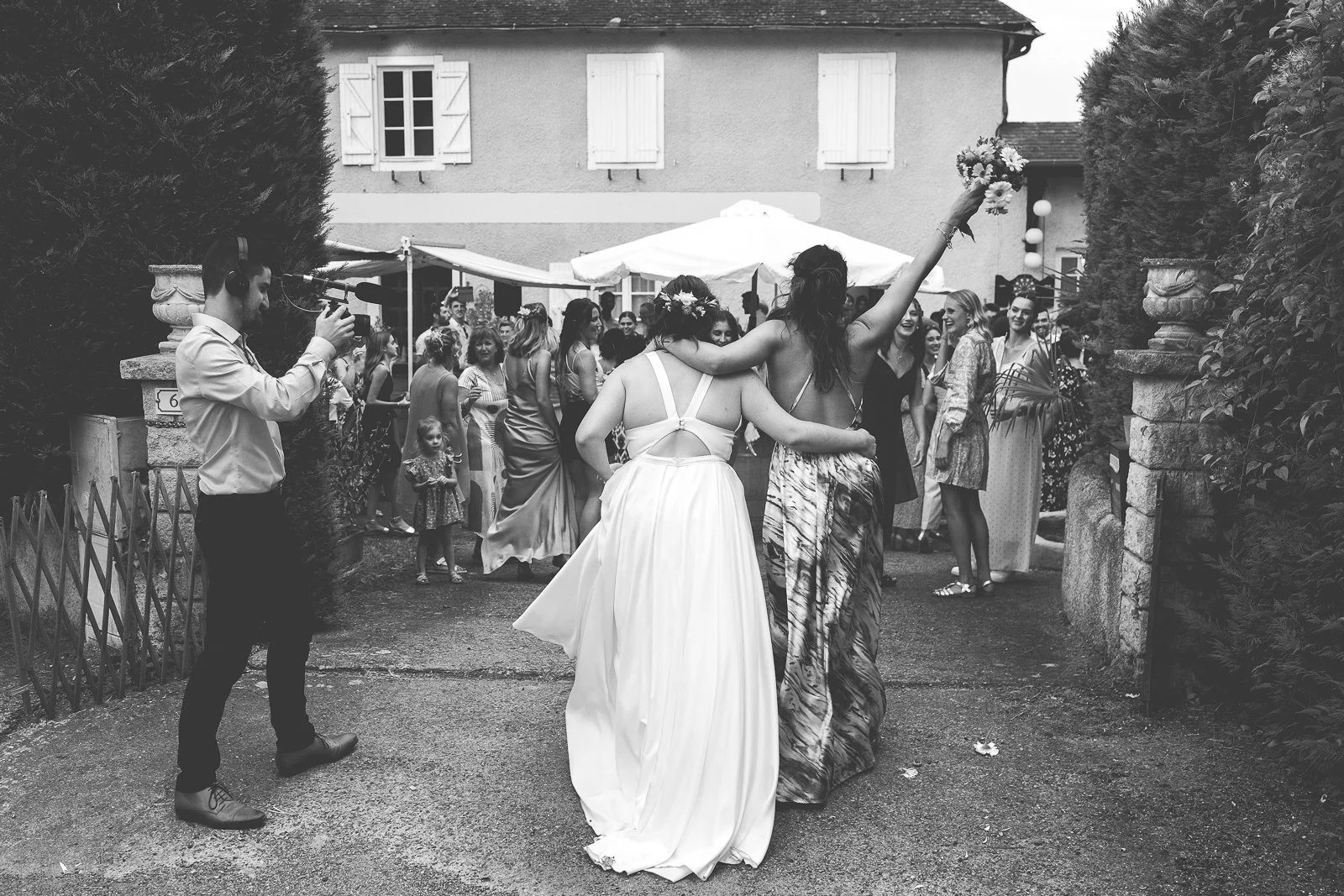 Une fête en extérieur avec un groupe de femmes qui dansent, dont deux portant des robes longues, une avec une robe blanche et une autre avec une robe à motif. Un homme filme la scène avec une caméra, alors que d’autres personnes regardent la danse, e