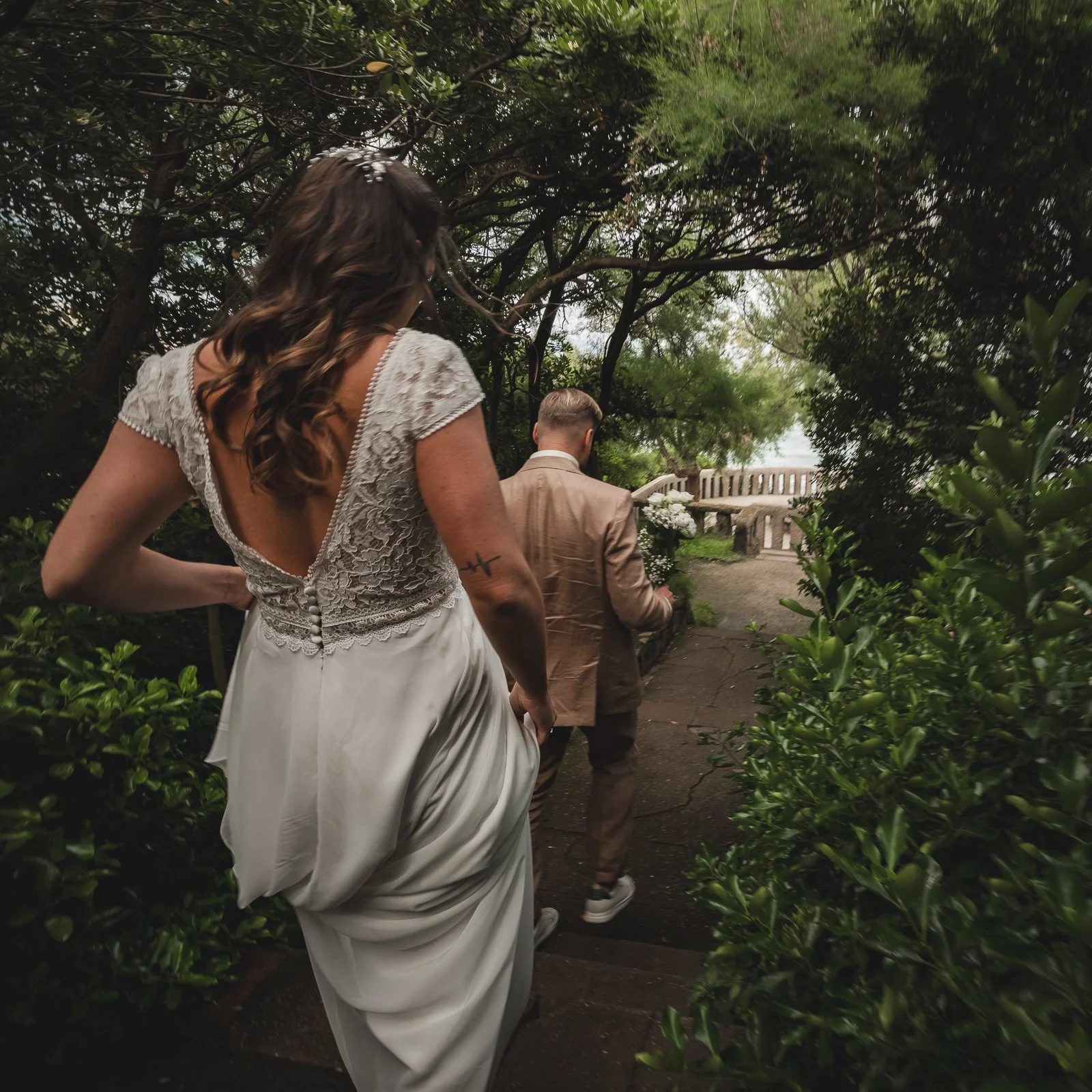 Un couple en mariage marche dans un jardin entouré de buissons et d'arbres, la femme porte une robe de mariage en dentelle et les cheveux bouclés, l'homme porte un costume beige.