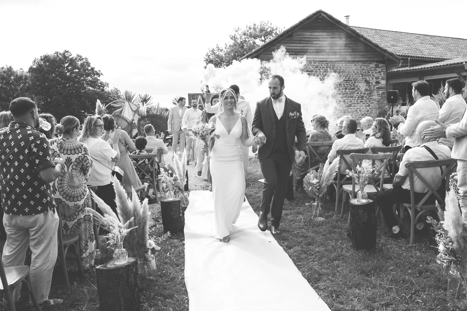 Couple marié marchant main dans la main lors d'une cérémonie de mariage en plein air, entourés de leurs invités, avec une demoiselle d'honneur portant un bouquet.