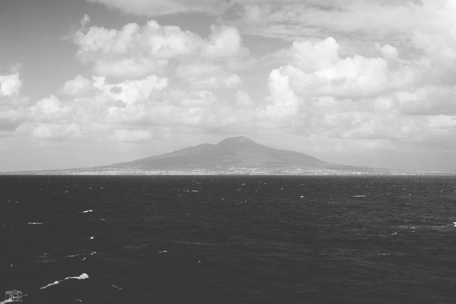 Vue d'une île volcanique au loin, probablement le mont Vésuve, vue depuis la mer avec des nuages dans le ciel.