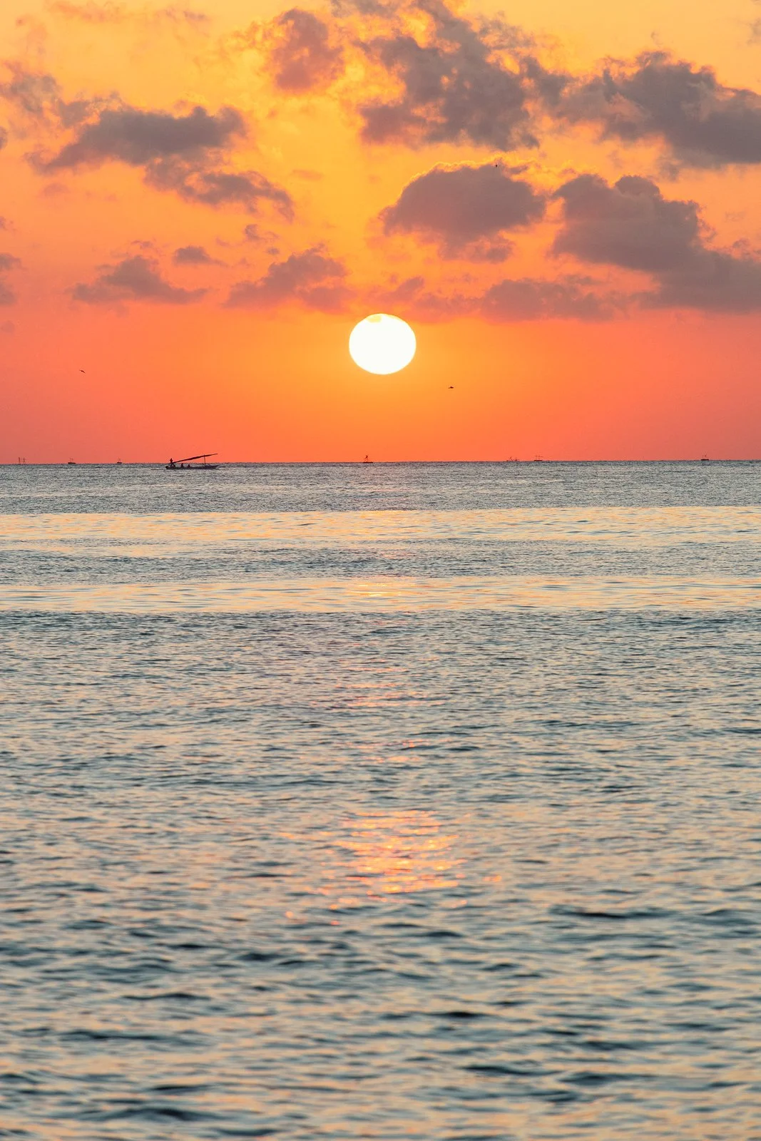 Coucher de soleil sur la mer avec un ciel orange et quelques nuages, bateau à voile en silhouette à l'horizon.