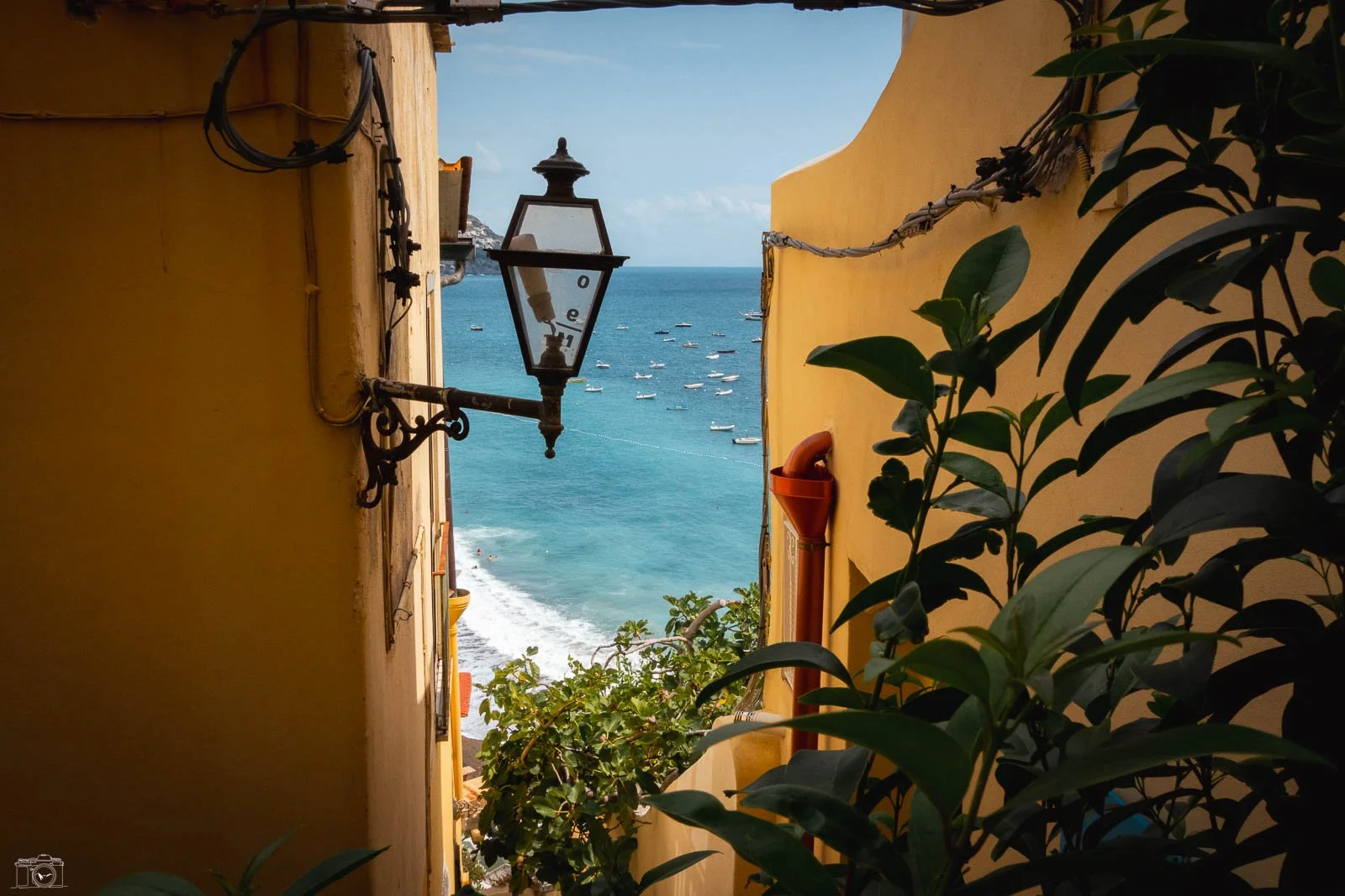 Vue d'une ruelle colorée menant à la mer, avec des bateaux au loin, une lampe ancienne suspendue entre deux murs jaunes, entourée de plantes vertes.