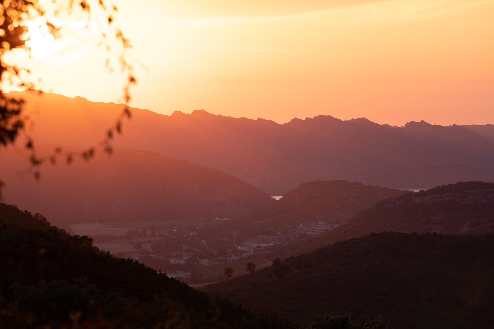 Paysage de montagnes au coucher du soleil avec un ciel orangé, vue sur une vallée et quelques bâtiments en contrebas.