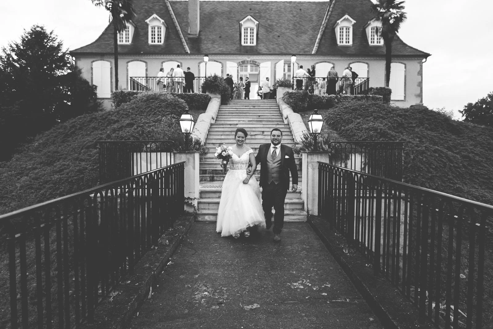 Un couple de mariés descend les marches d'une grande maison lors d'un mariage, entourés de invités au second étage de la maison, dans un cadre extérieur avec des buissons et des lampadaires.