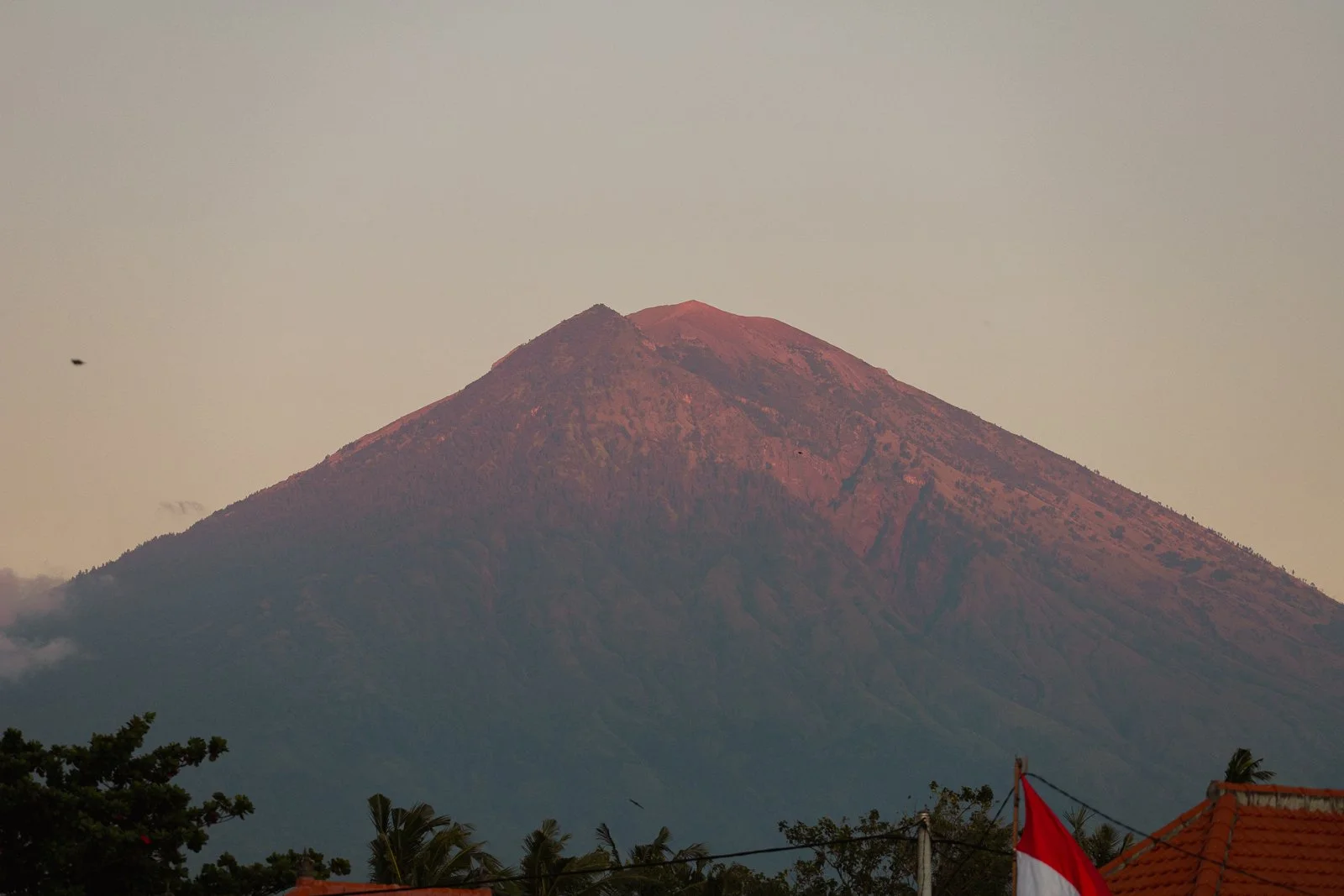 Montagne volcanique avec sommet rougeâtre au coucher du soleil, avec quelques arbres et bâtiments au premier plan.