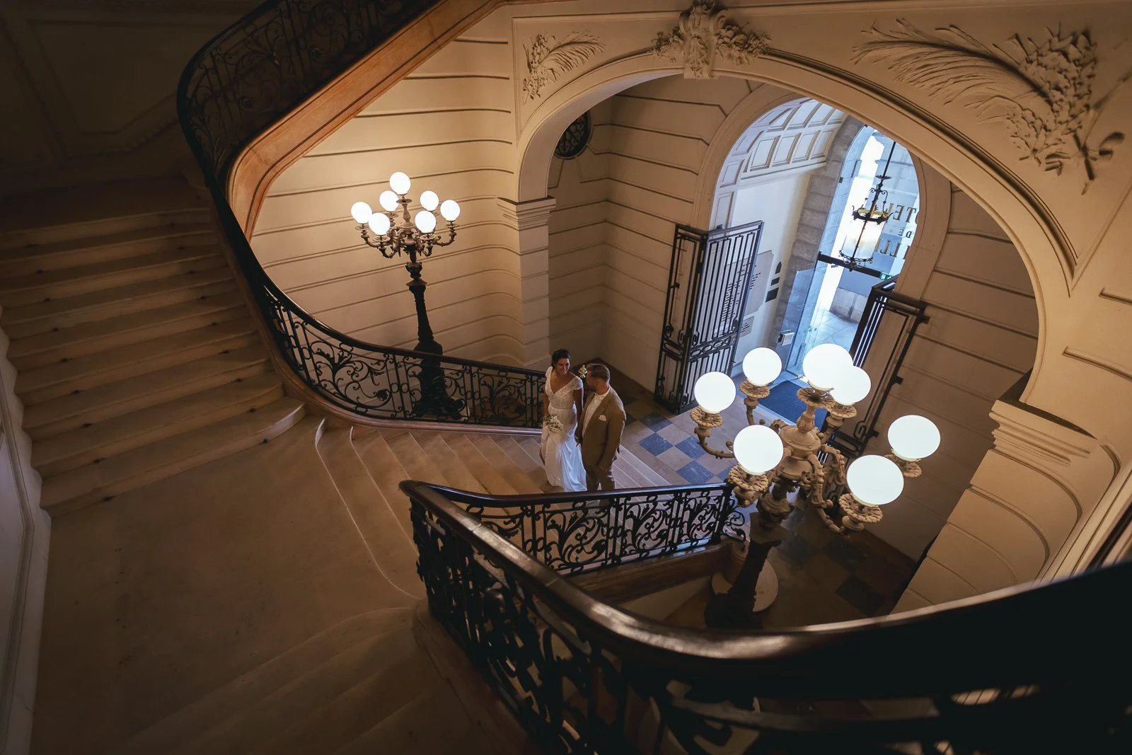 Un couple de mariés se tient dans une entrée élégante, au centre de l'escalier en colimaçon avec des balustrades en fer forgé, entouré d'anciens luminaires à globes lumineux dans un bâtiment historique.