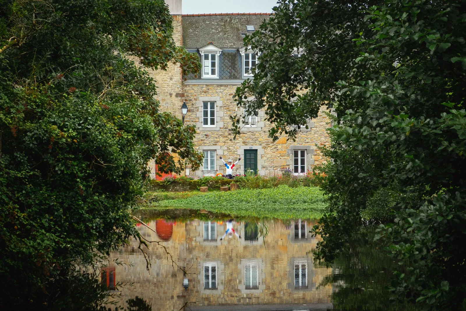 Une maison en pierre vue à travers des arbres, avec un reflet dans l'eau en première plan, présence d'une sculpture de cycliste au centre.