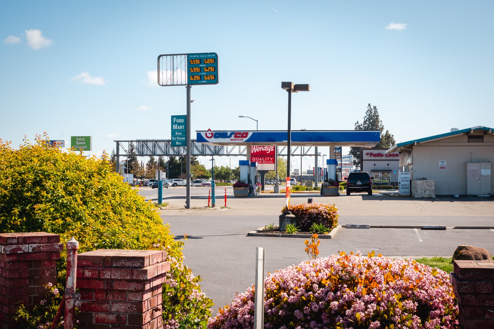 Station-service avec panneaux indiquant le prix de l'essence, commerces environnants, fleurs et arbustes en premier plan.