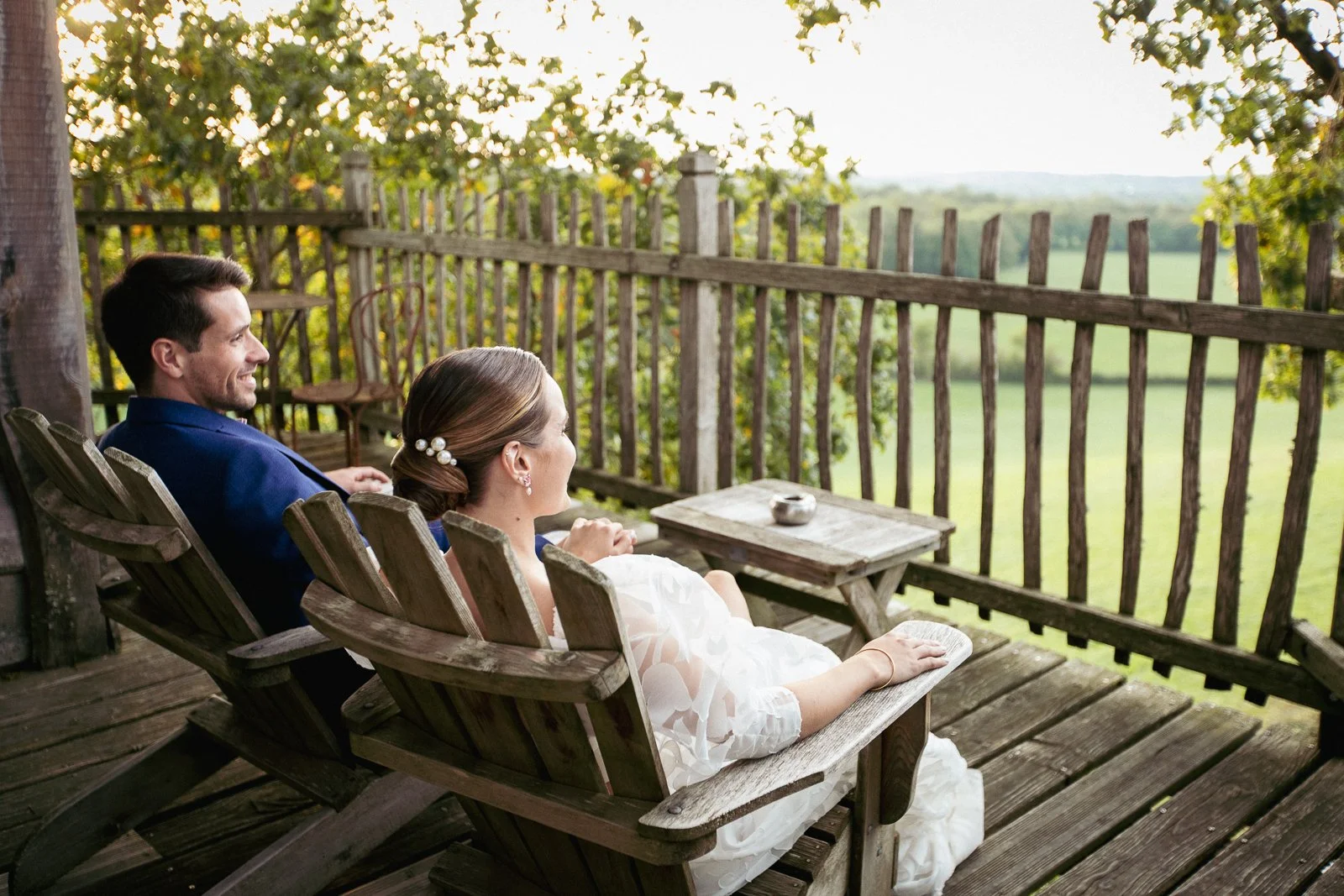 Un couple en robe de mariée et costume assis sur un balcon en bois, regardant un paysage verdoyant relaxant, lors d'une journée ensoleillée.