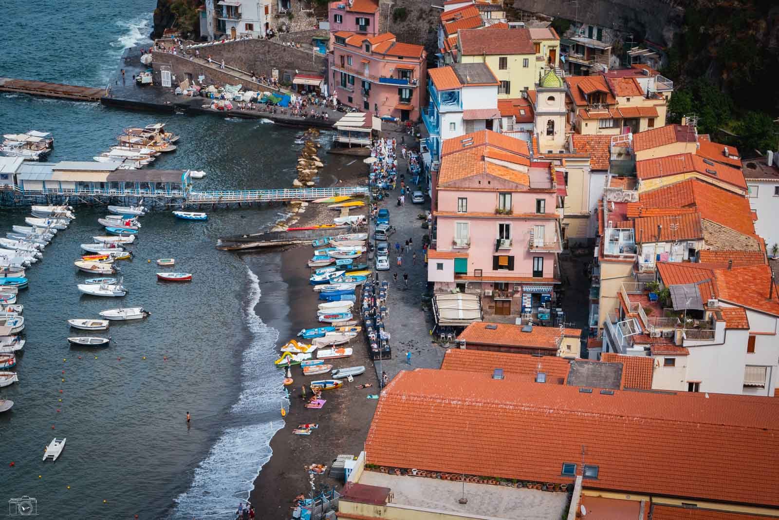 Vues aériennes d'une côte italienne avec un port rempli de petits bateaux, des bâtiments colorés avec toits en tuiles rouges, et une foule de personnes sur la plage et la promenade.