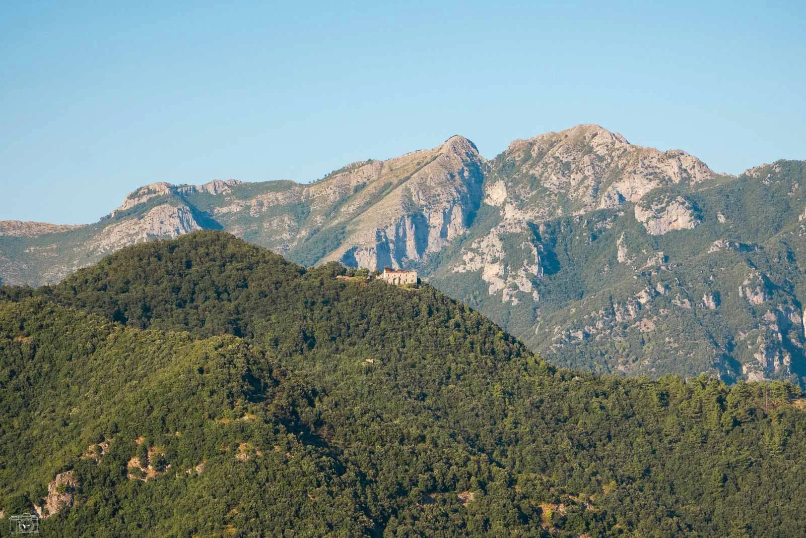 Montagnes boisées avec un petit bâtiment sur une colline en premier plan et des montagnes rocheuses en arrière-plan.