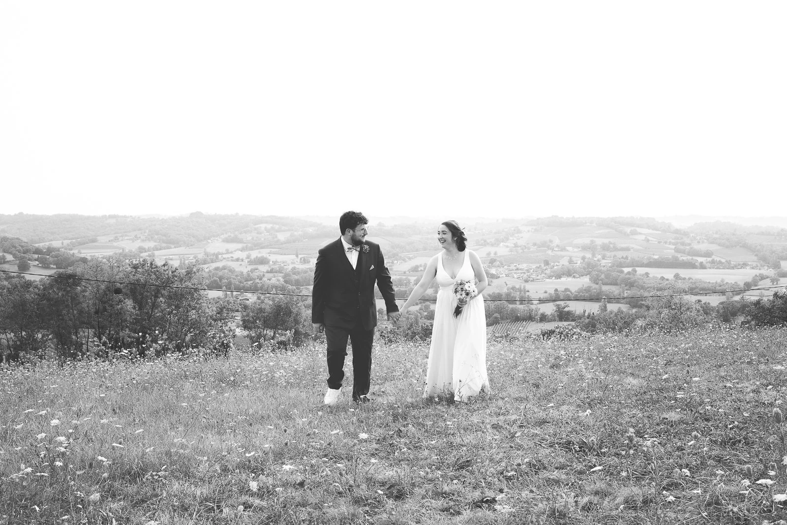 Un couple de mariés marche main dans la main dans un champ avec un paysage rural en arrière-plan. La femme porte une robe blanche et tient un bouquet, l'homme porte un costume noir.