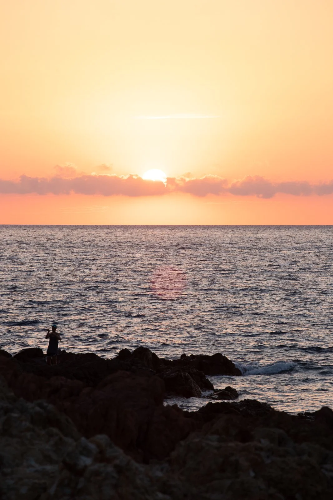 Un coucher de soleil sur l'océan avec un ciel orangé et une personne debout sur les rochers en regardant la mer.