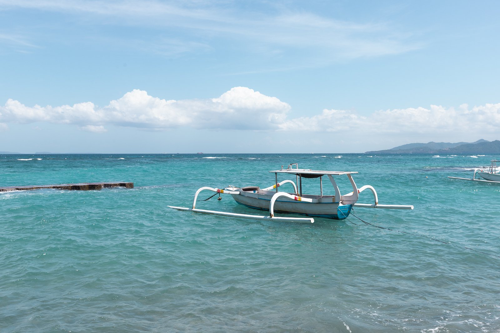 Un bateau traditionnel en bois avec des rameurs en métal flottant sur une mer turquoise sous un ciel bleu avec quelques nuages, et une montagne en arrière-plan.