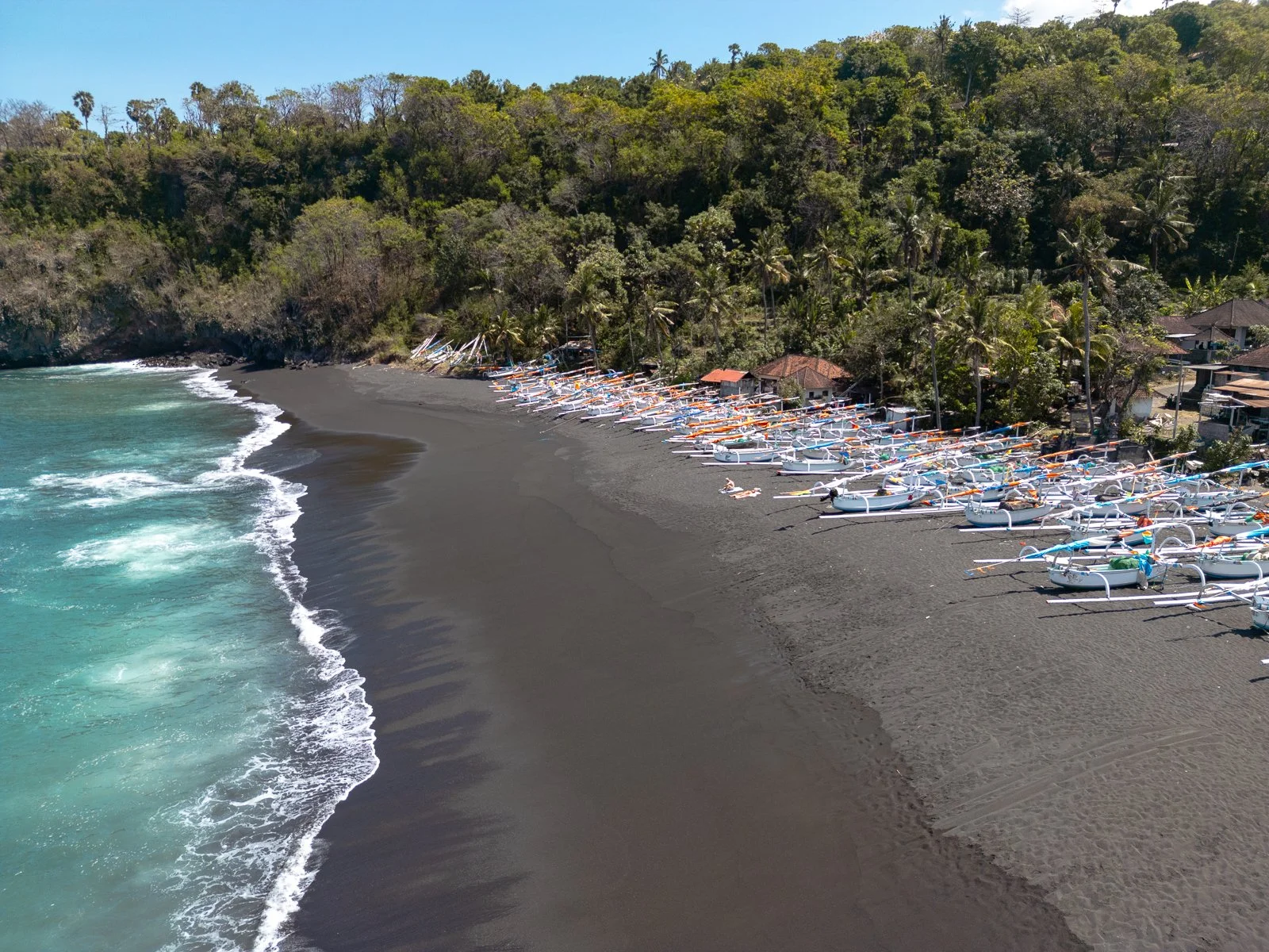 Une plage de sable noir avec plusieurs bateaux de pêche traditionnels alignés sur le rivage et une végétation dense en arrière-plan.