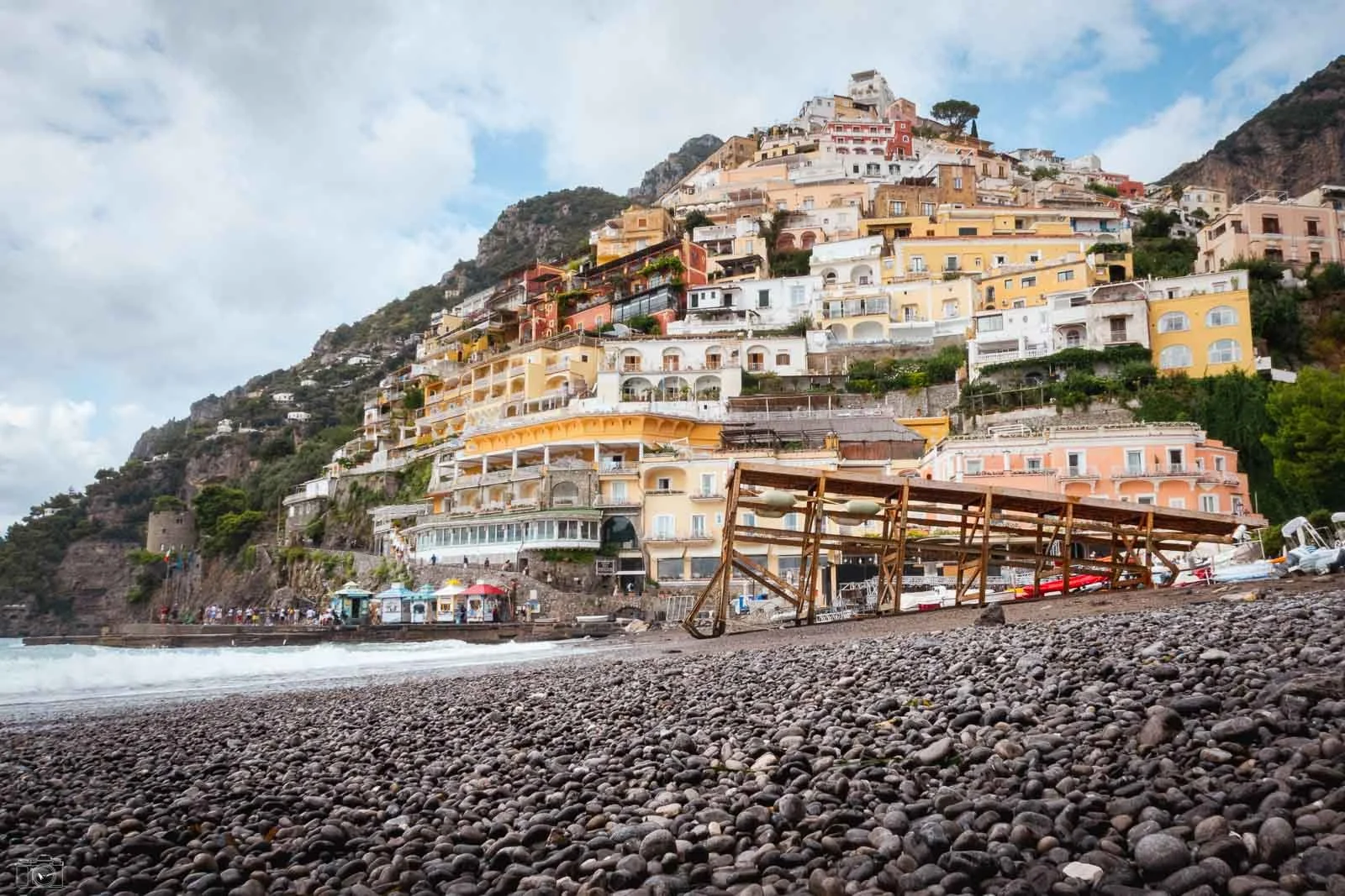 Bâtiments colorés construits en terrasses sur une colline escarpée, vue depuis un rivage rocheux avec un bateau en bois en premier plan.
