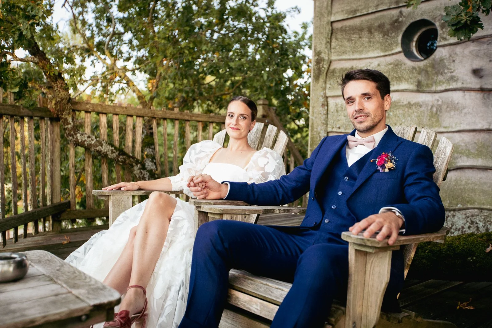 Un couple de mariés assis sur des chaises en bois dans une terrasse en extérieur, tenant la main, lors d'un mariage