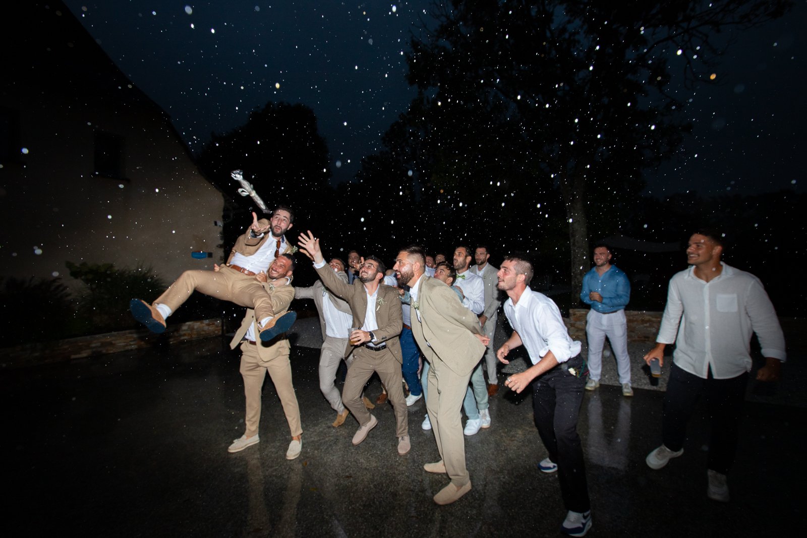 Un groupe d'hommes habillés en costumes fête lors d'une soirée en plein air sous la pluie, certains rient et d'autres dansent.