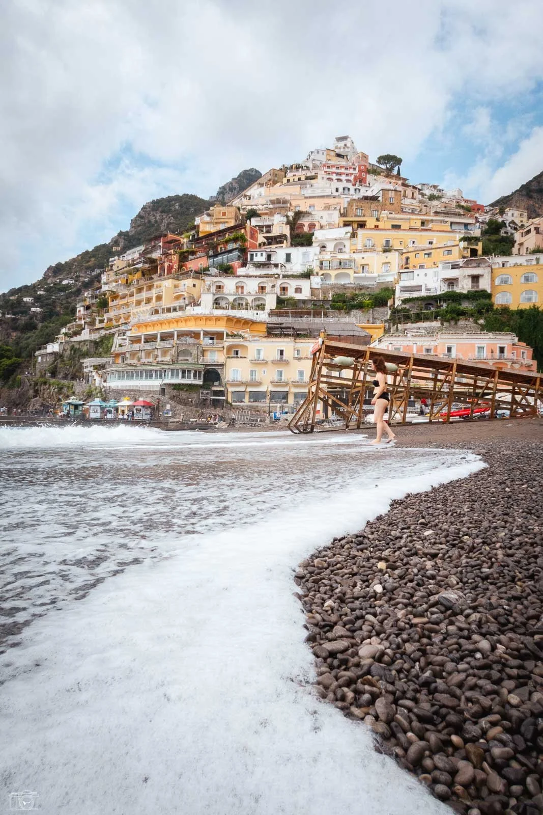 Un paysage côtier avec une plage de galets, une femme en maillot de bain se tenant à côté d'une structure en bois, et une ville colorée perchée en haut d'une colline, avec des bâtiments multicolores et une montagne en arrière-plan.
