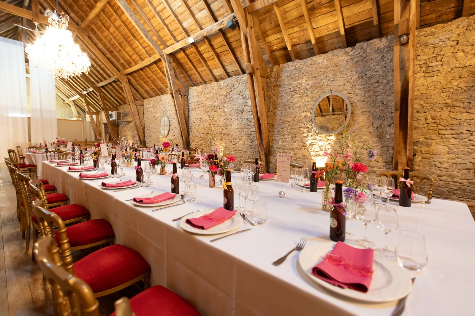Salle de fête avec une longue table dressée pour un événement, décorée avec des fleurs, des bouteilles de bière, des verres à vin et des serviettes roses, dans un lieu avec des murs en pierre et un plafond en bois.