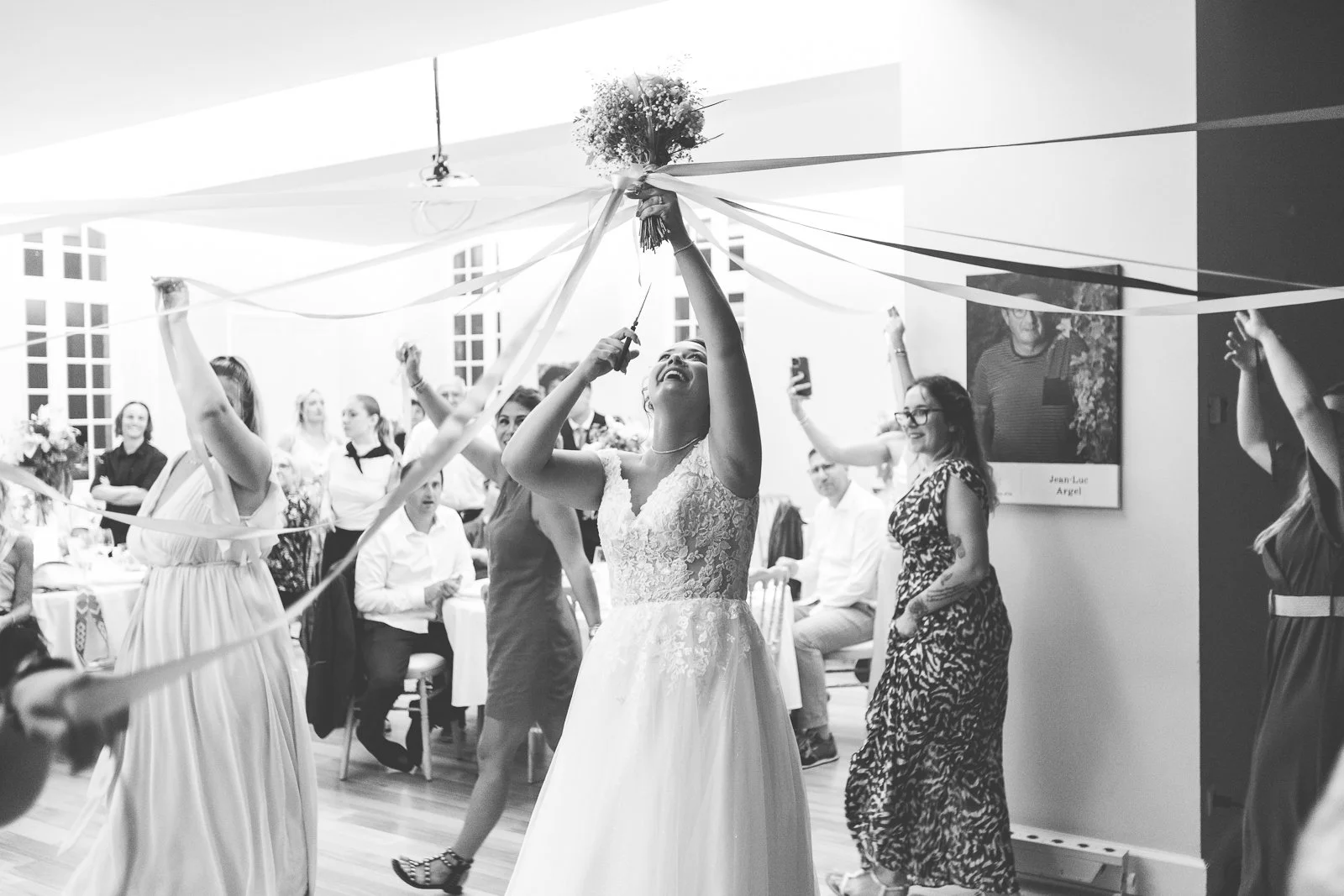 Une femme avec une robe de mariage en train de couper un bouquet lors d'une fête ou d'une célébration, entourée de plusieurs personnes qui dansent et prennent des photos. La scène est en noir et blanc.