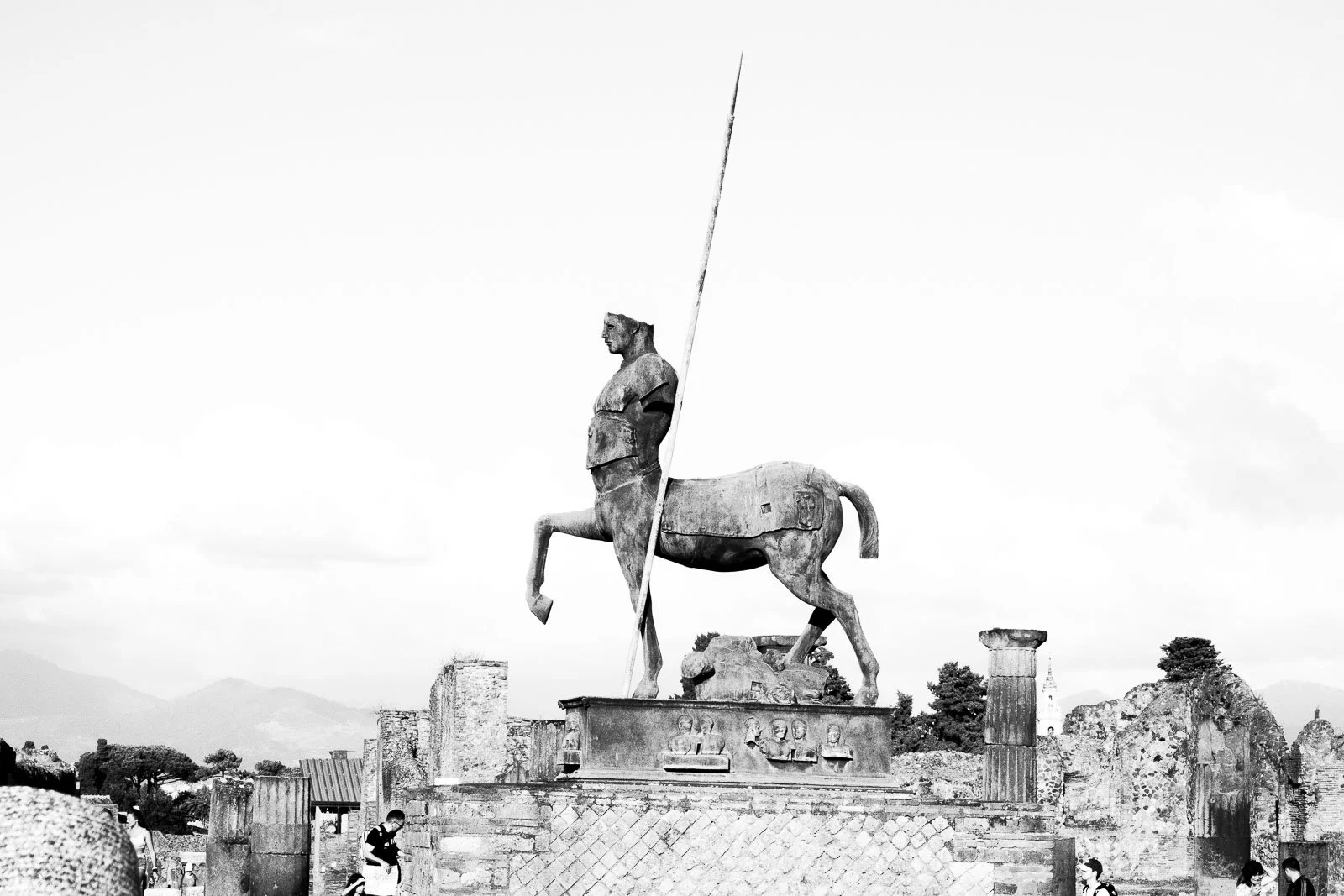 Statue en bronze d'un centaure avec une figure humaine à la tête d'enfant, située en plein air, avec des ruines antiques et des montagnes en arrière-plan, et des visiteurs devant la statue.