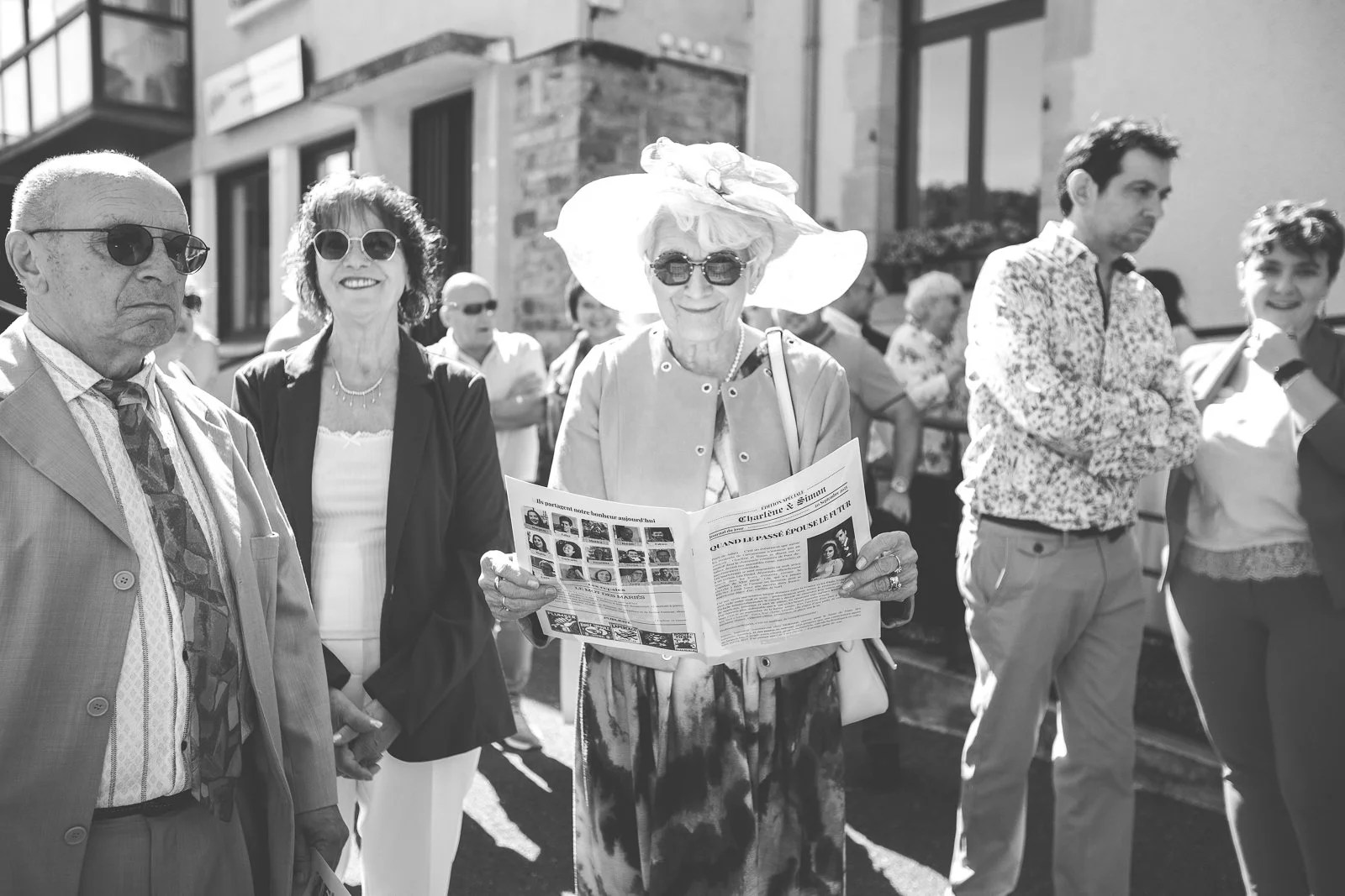Groupe de personnes en plein air, dont une femme âgée portant un grand chapeau et des lunettes, lisant un journal. Autres personnes autour, portant des lunettes, dans un contexte ensoleillé.