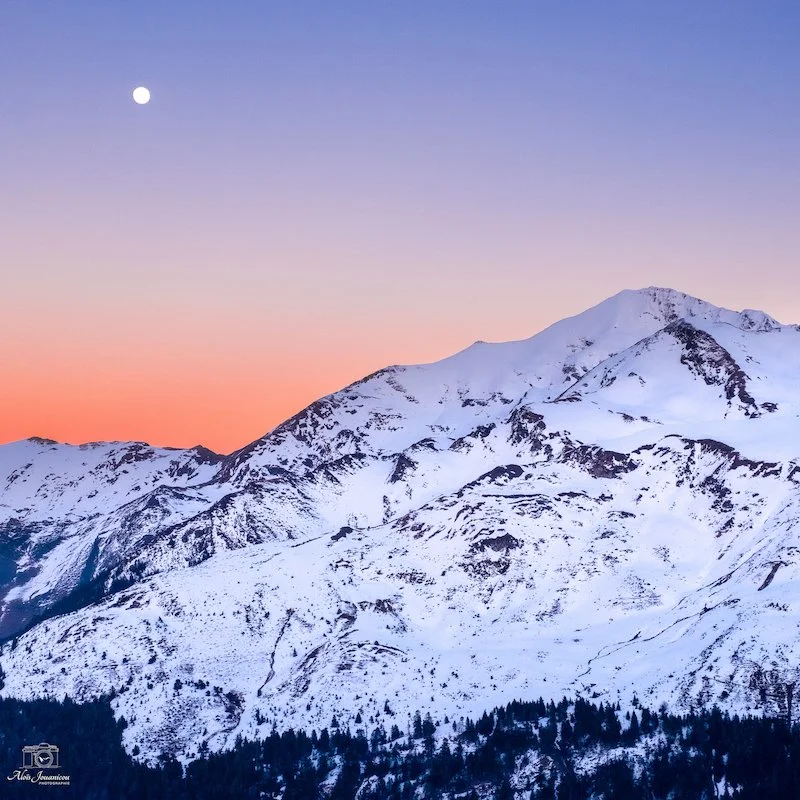Montagnes enneigées au coucher du soleil avec un ciel coloré et la lune visible.